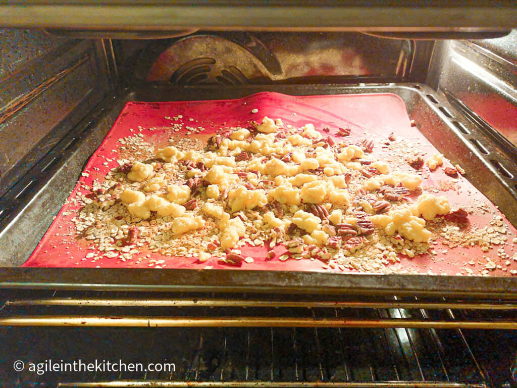 A photo taken from the oven on a cookie sheet, a red silicone sheet with oats, cinnamon, pecans and cookie crumbles baking.