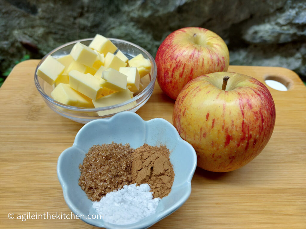 On a cutting board, the ingredients to make fragrant baked apple, from top left, a bowl of cubed butter, two red and yellow apples, a small bowl of brown sugar, cinnamon and vanilla sugar