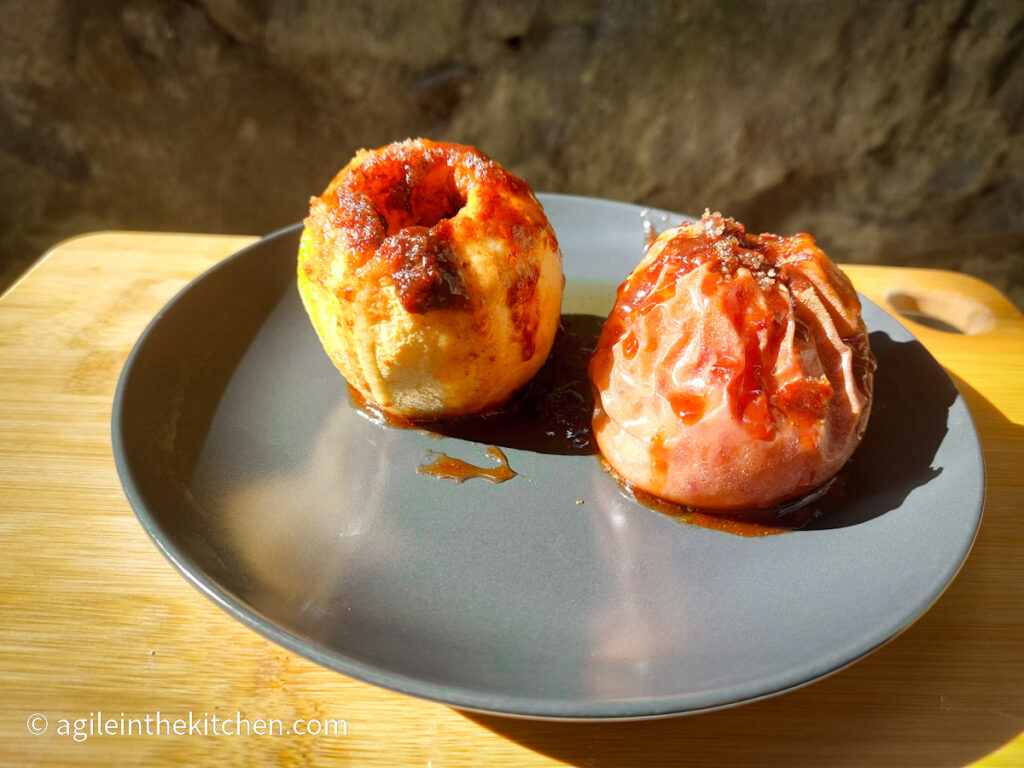 On a grey plate, two baked apples with a cinnamon mixture, one peeled, one unpeeled.
