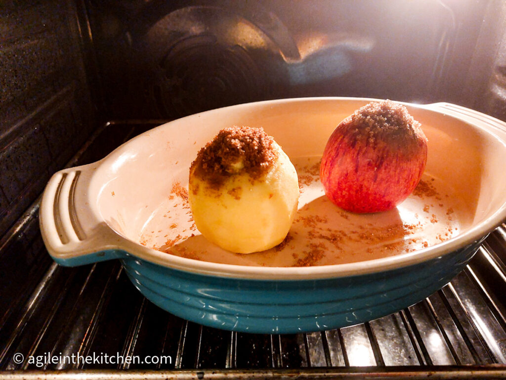 A photo taken from the oven, a blue oven proofed dish with two baked apples inside, one peeled, one unpeeled with cinnamon mixture coming out of the core.