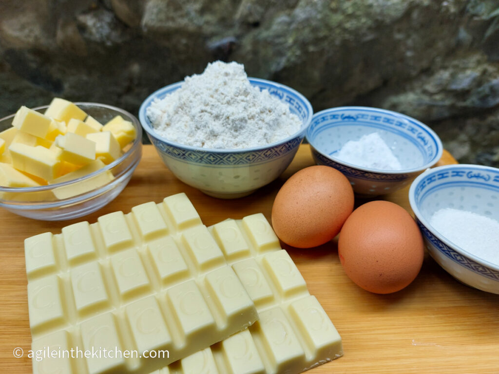 On a wooden cutting board, ingredients to make white chocolate cake, from top left, a bowl of cubed butter, a bowl of flour, a bowl of vanilla sugar, a bowl of white sugar, two eggs and white chocolate bars.