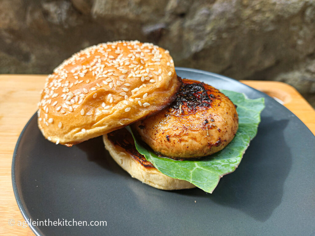 On a wooden cutting board a black matte plate with a brioche hamburger bun with sesame seeds, a marinated mushroom burger and a sheet of leaf lettuce.