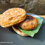 On a wooden cutting board a black matte plate with a brioche hamburger bun with sesame seeds, a marinated mushroom burger and a sheet of leaf lettuce.