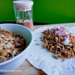 On a cutting board, in the foreground to the left a bowl of shredded tofu taco. In the background a jar of home made tacoseasoning. To the left a flour tortilla with shredded tofu taco with chopped red onions on top.