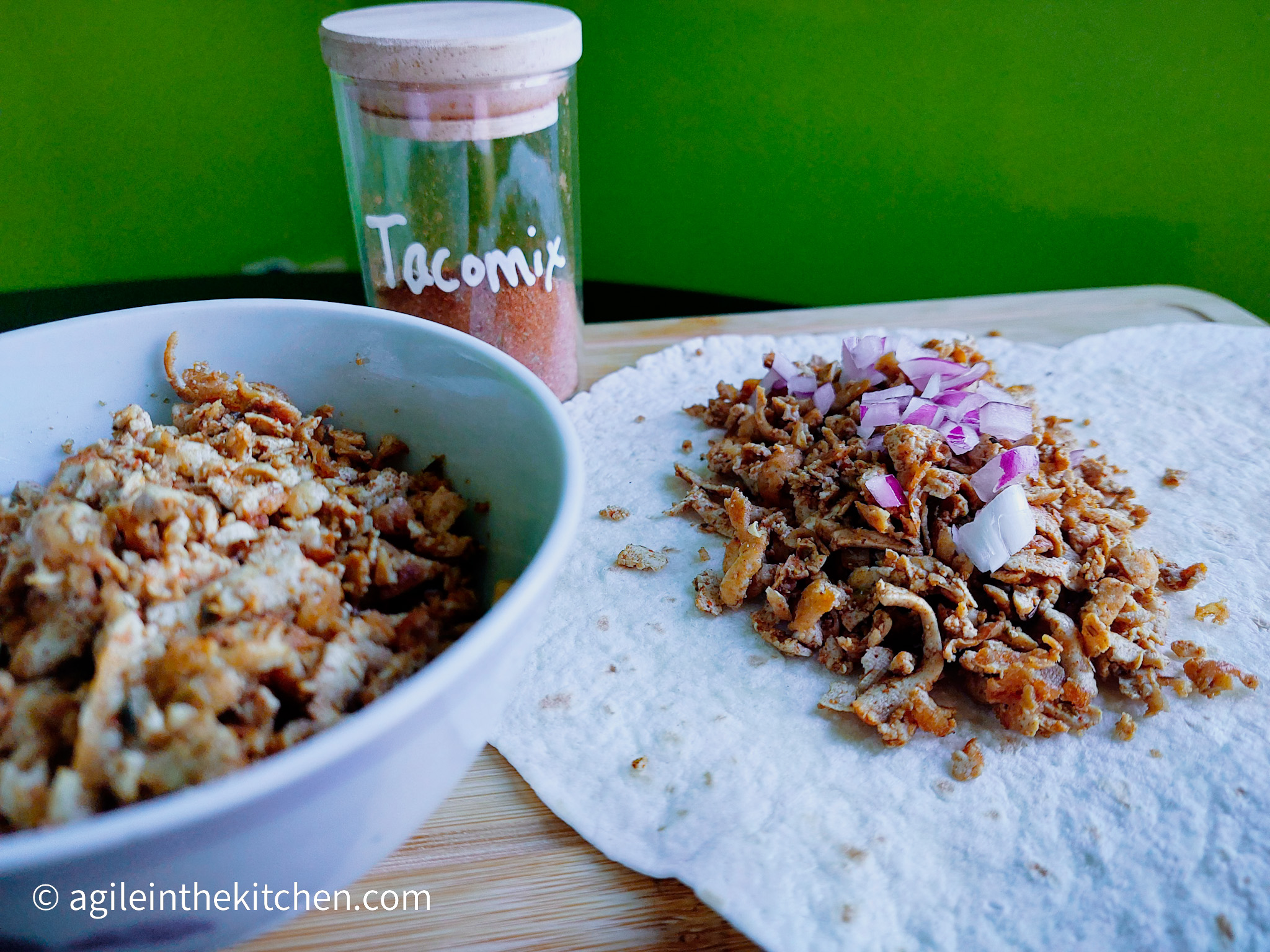 On a cutting board, in the foreground to the left a bowl of shredded tofu taco. In the background a jar of home made tacoseasoning. To the left a flour tortilla with shredded tofu taco with chopped red onions on top.