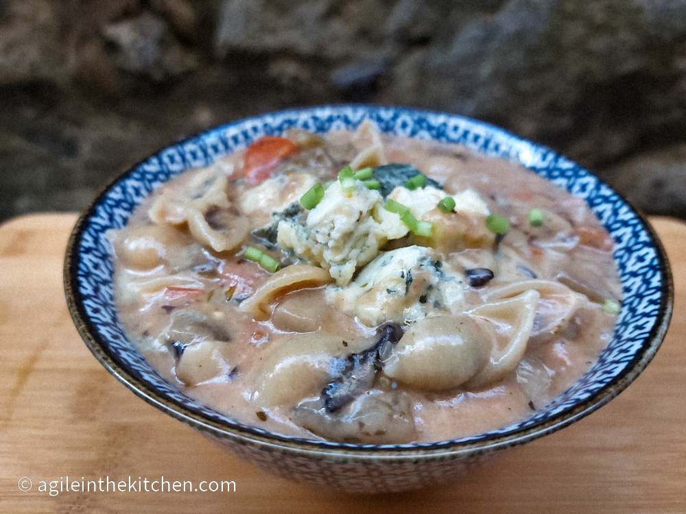 On a wooden cutting board, a blue ceramic bowl filled with bluecheese pasta topped with green onions