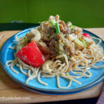On a wooden cuttingboard with a pink cloth napkin on the right, a blue plate topped with vegetarian fakeaway stirfry with noodles, brockli, red bellpeppers, cashew nuts and topped with green onion