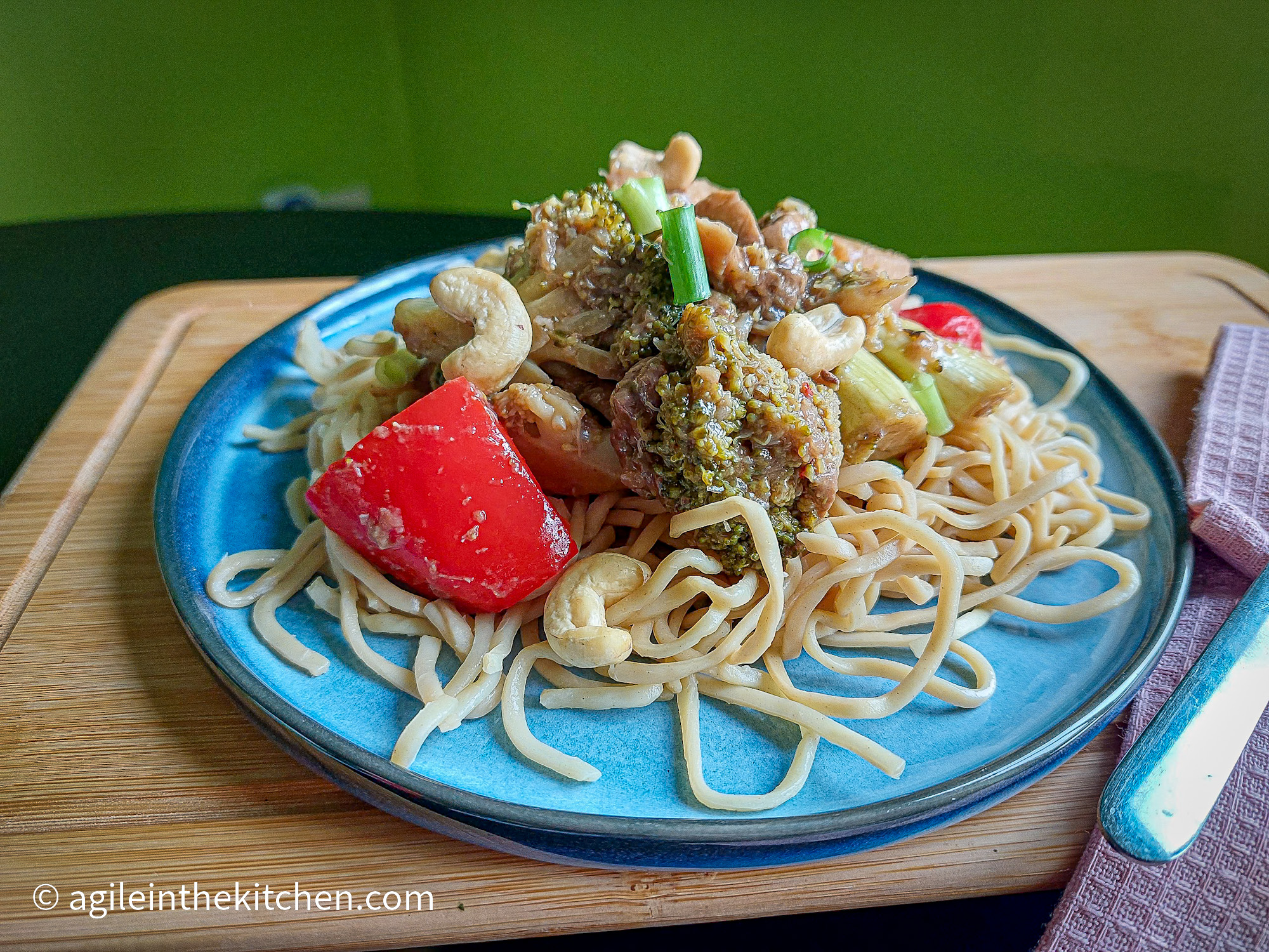 On a wooden cuttingboard with a pink cloth napkin on the right, a blue plate topped with vegetarian fakeaway stirfry with noodles, brockli, red bellpeppers, cashew nuts and topped with green onion