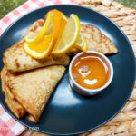 A black plate with pancakes folded with orange and lemon slices, next to them a small pot of citrus syrup