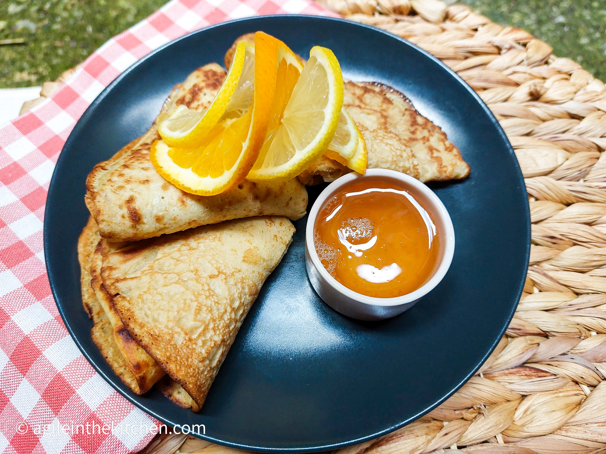 A black plate with pancakes folded with orange and lemon slices, next to them a small pot of citrus syrup