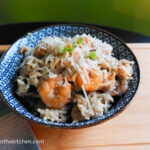 On a wooden cutting board, a blue patterned ceramic bowl with rice and shrimp, topped with shredded parmesan and chopped green onions. To the left is a blue, textured cloth napkin.
