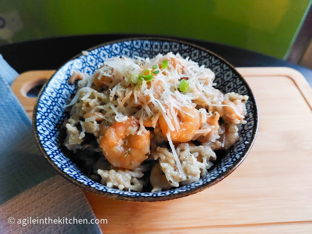 On a wooden cutting board, a blue patterned ceramic bowl with rice and shrimp, topped with shredded parmesan and chopped green onions. To the left is a blue, textured cloth napkin.