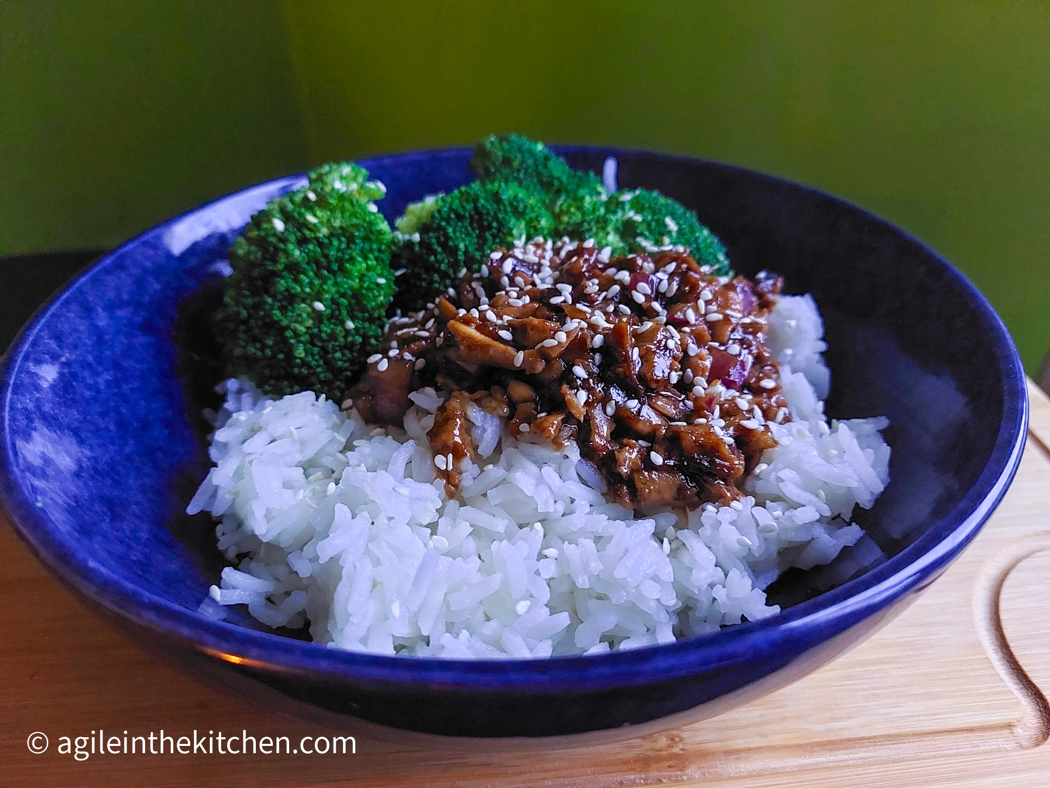 On a wooden cutting board a blue bowl with rice topped with shredded teriyaki tofu, broccoli and a sprinkling of sesame seeds