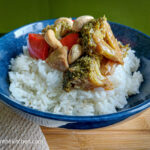 On a wooden cutting board a blue bowl with rice and vegan stir fry with broccoli, red bell peppers and cashew nuts