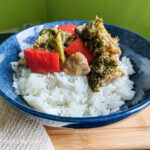 On a wooden cutting board with a beige cloth napkin on the left, a darkblue shallow bowl with rice topped with vegan stirfry fakeaway, with brockoli, muhsroom, red bell peppers and cashew nuts.