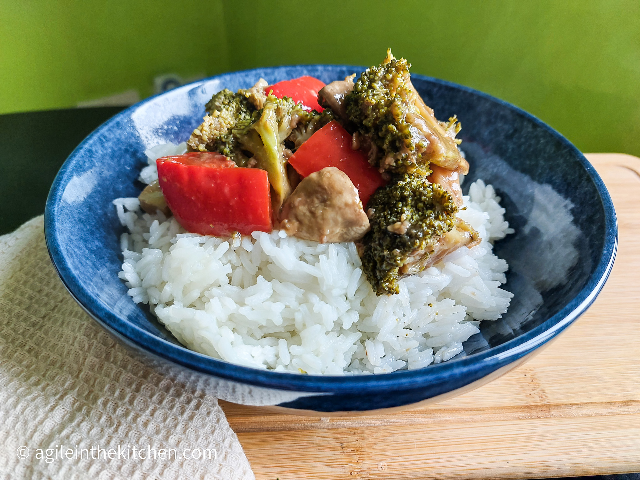 On a wooden cutting board with a beige cloth napkin on the left, a darkblue shallow bowl with rice topped with vegan stirfry fakeaway, with brockoli, muhsroom, red bell peppers and cashew nuts.
