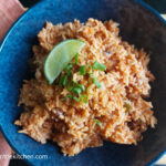 A blue shallow bowl with vegan rice and bean, a wedge of lime and cut green onions on top. To the left a folded pink cloth napkin