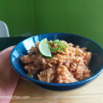 On a wooden cutting board a blue shallow bowl with vegan rice and bean, a wedge of lime and cut green onions on top. To the left a folded pink cloth napkin