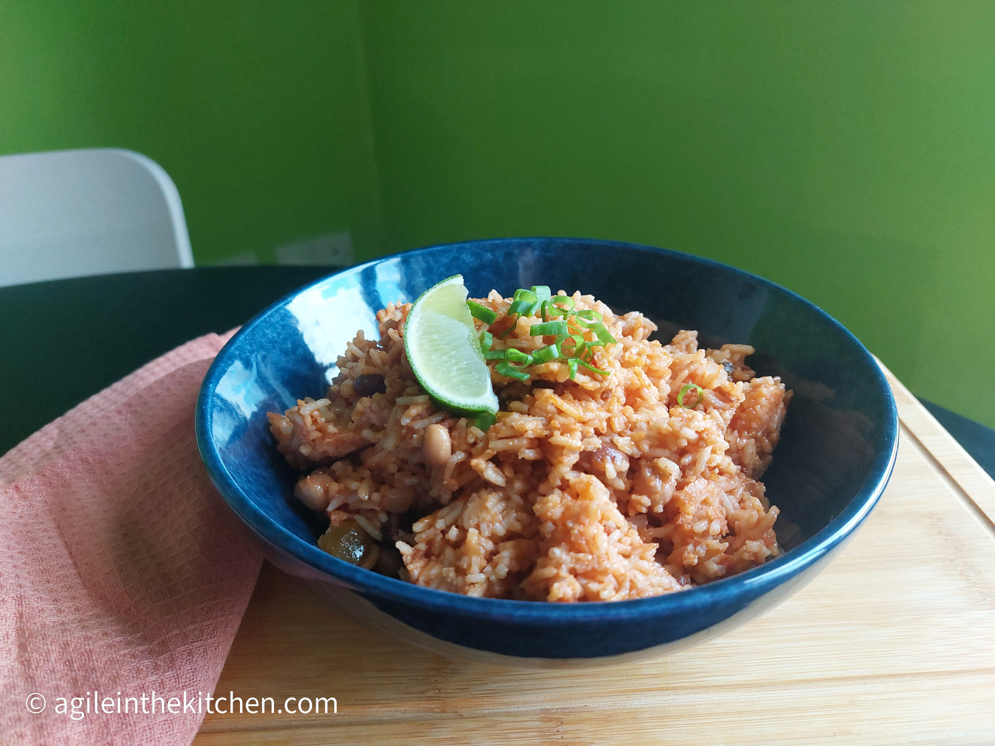On a wooden cutting board a blue shallow bowl with vegan rice and bean, a wedge of lime and cut green onions on top. To the left a folded pink cloth napkin