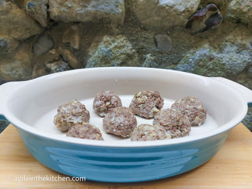 On a cutting board with a stone wall background, a blue Le Creuset oven pan with eight asian meatballs