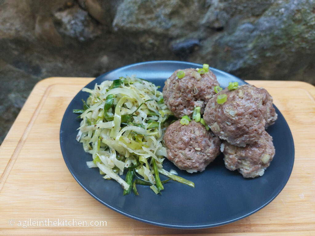 On a wooden cutting board with a stone wall background a black matte plate with Asian inspired meatballs pilled up to the right and to the left a handfull of pickled Irish cabbage salad.
