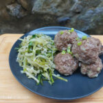 On a wooden cutting board with a stone wall background a black matte plate with Asian inspired meatballs pilled up to the right and to the left a handfull of pickled Irish cabbage salad.