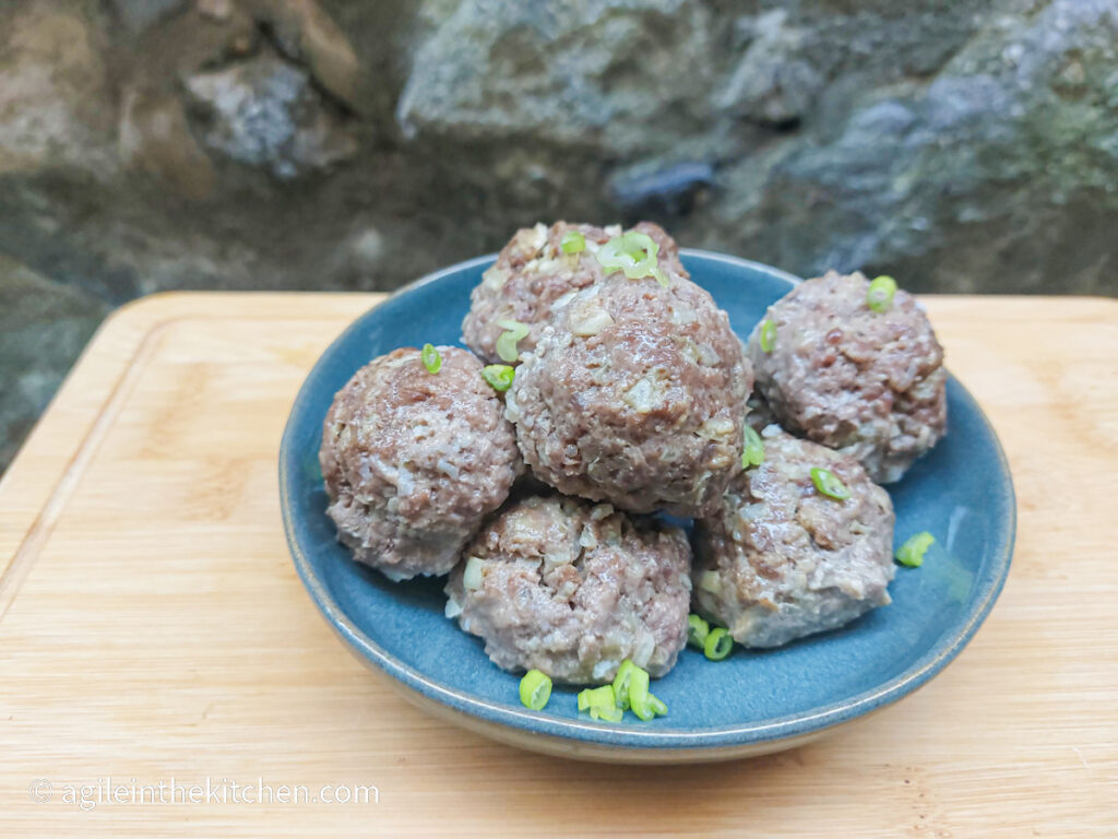 On a wooden cutting board with a stone wall background a blue plate with Asian inspired meatballs pilled up with a sprinkling of green onions.