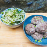 On a wooden cutting board with a stone background a bowl with pickled cabbage salad and a blue plate with six asian meatballs, sprinkled with green onions.