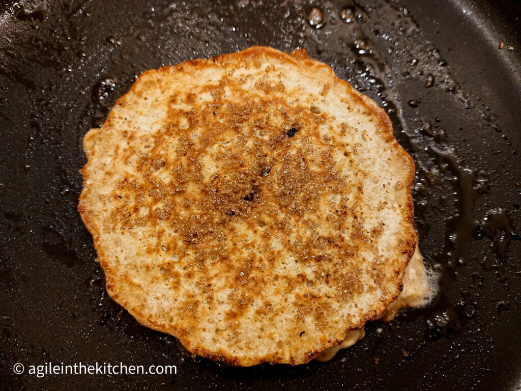In a frying pan, a pancake s being fried in butter, with a sprinkle of brown sugar on top of the batter.