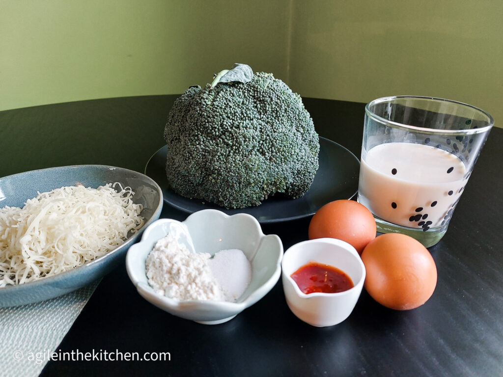 On a black table, ingredients to make broccoli pancake. From left a plate of shredded cheese, a head of broccoli, a flower shaped bowl with bakingpowder and salt, a small bowl of sambal olek, two eggs and a glass of milk