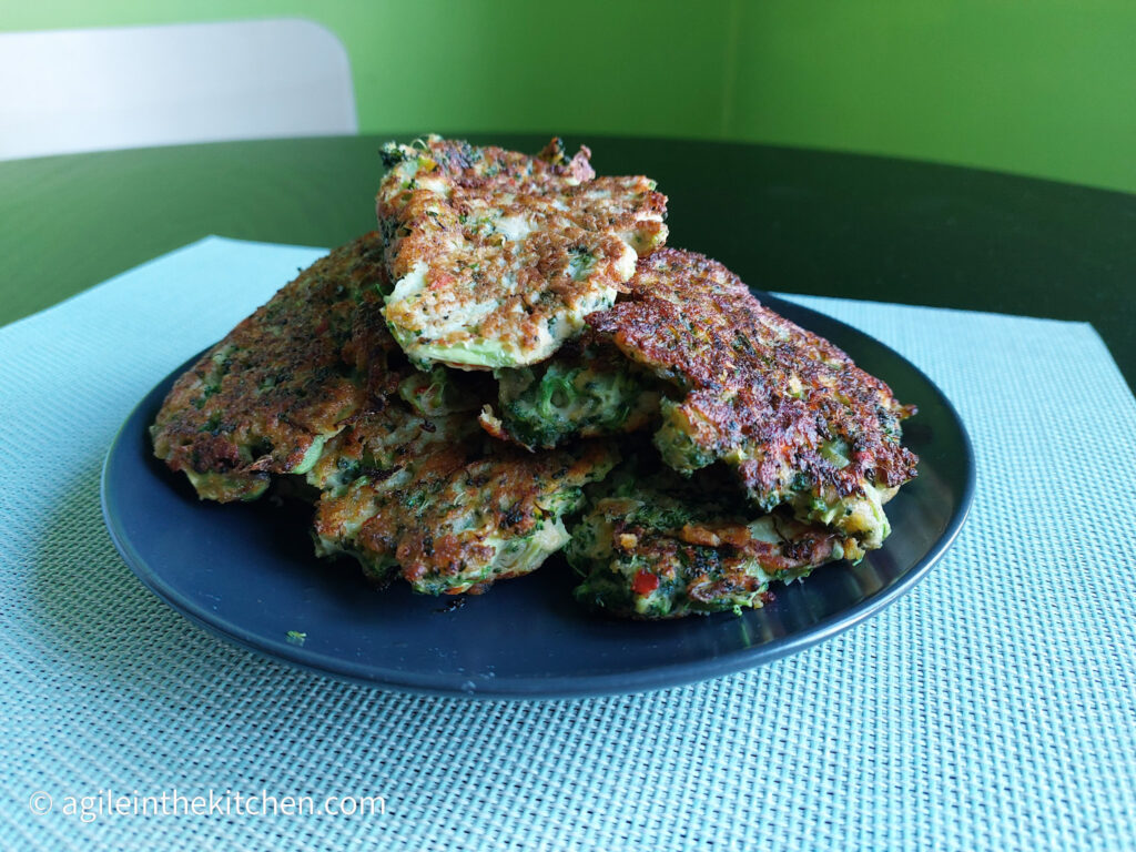 On a blue textured placemat, a black matte plate, piled high with broccoli pancakes